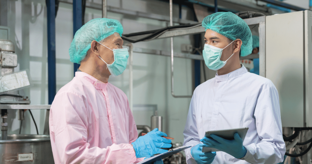 Two capsule filling machine operators wearing PPE, holding clipboards, and performing quality assurance testing.