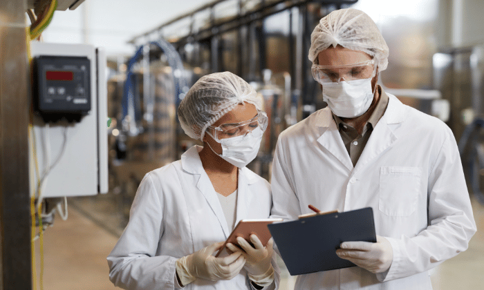 Two lab-coated operators in hairnets and masks reviewing plans in a manufacturing facility for capsule filling production.
