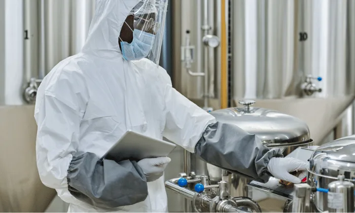 Technician in full protective gear inspecting and adjusting stainless steel pharmaceutical equipment in a cleanroom environment.