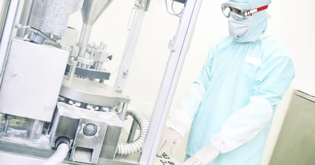 An operator standing in front of a capsule filling machine wearing PPE such as gloves, goggles, and a mask.